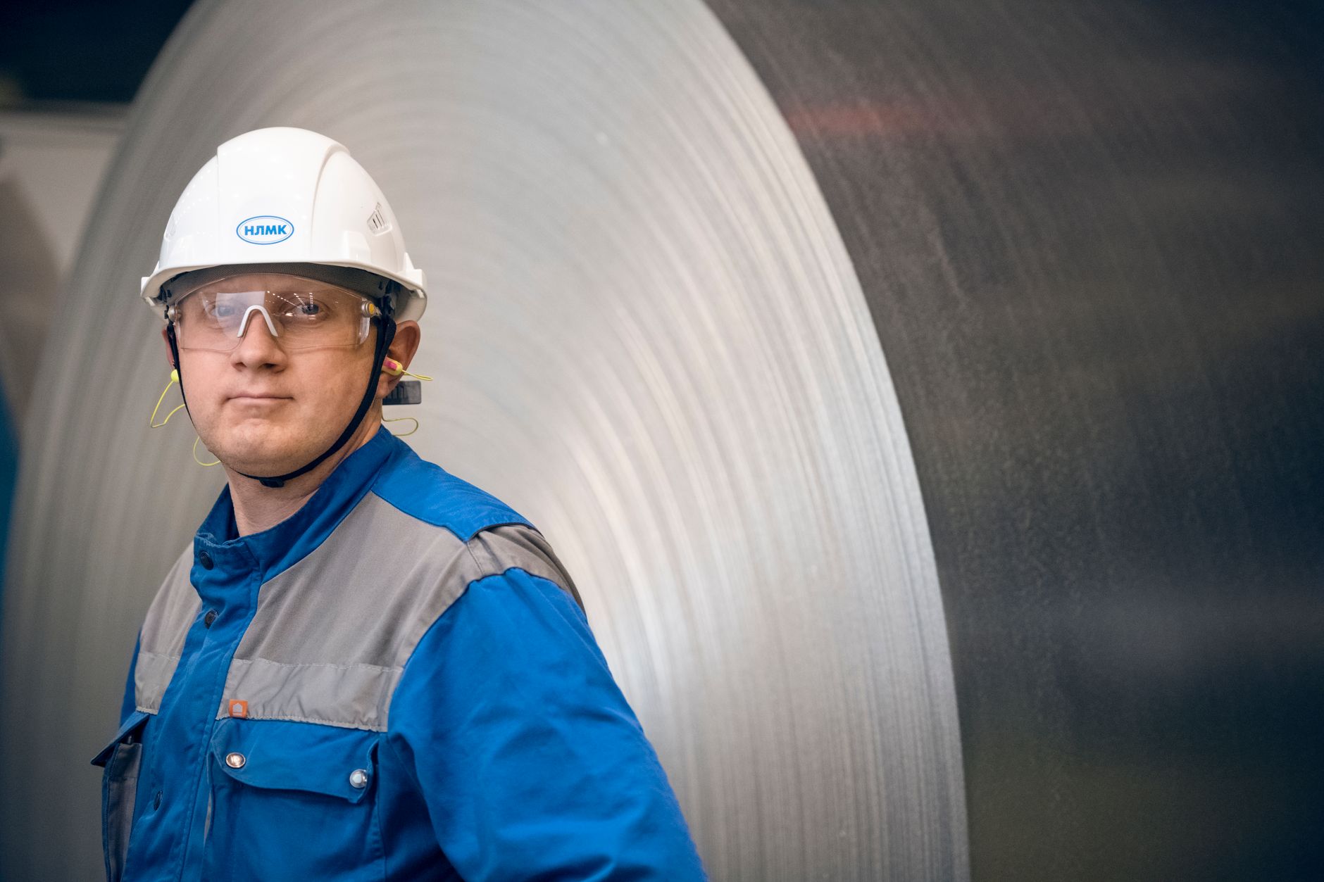 Portrait of a factory worker in protective gear in front of a large steel coil in an industrial setting.