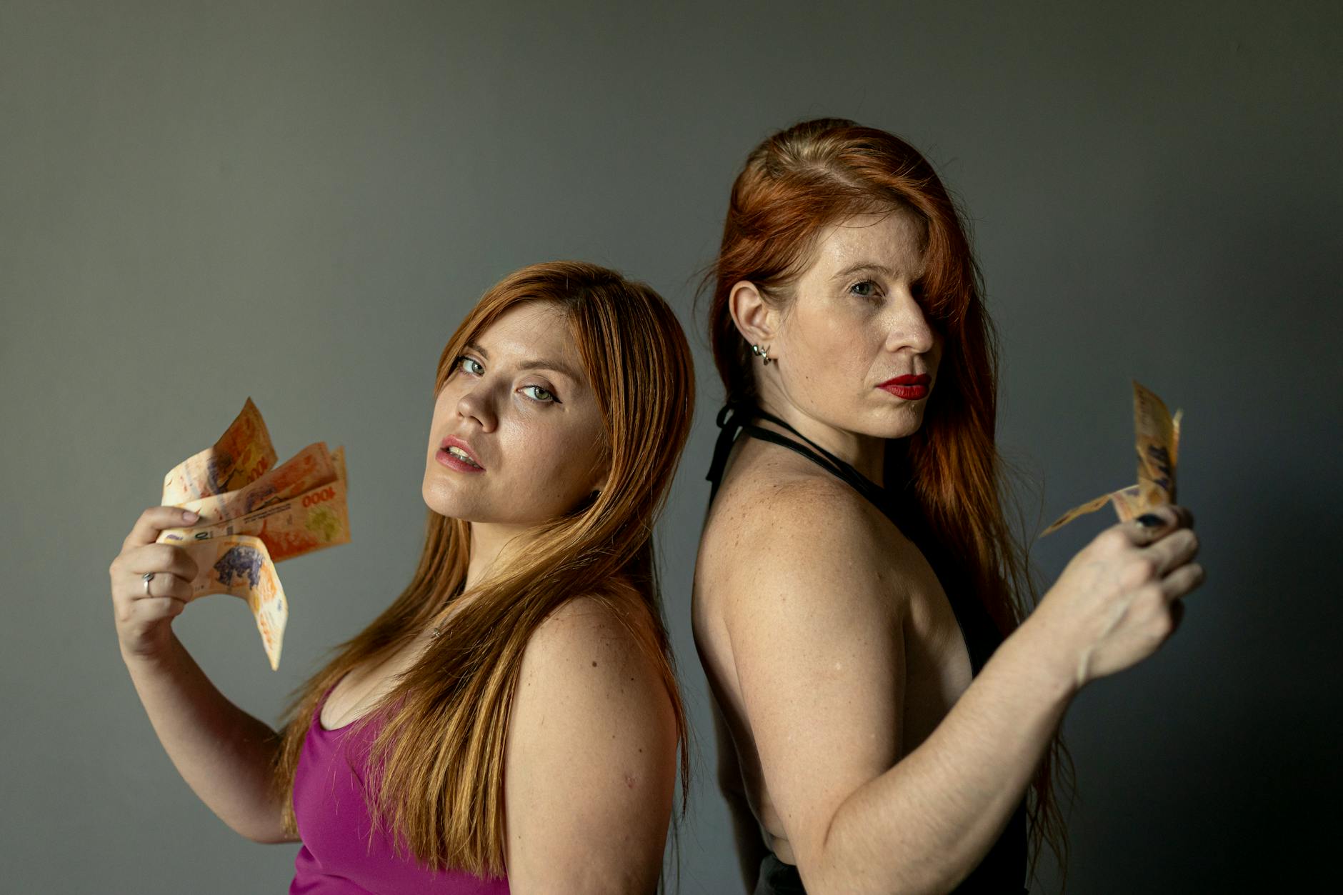 Portrait of two women displaying Argentine pesos. Shot indoors with a confident look.