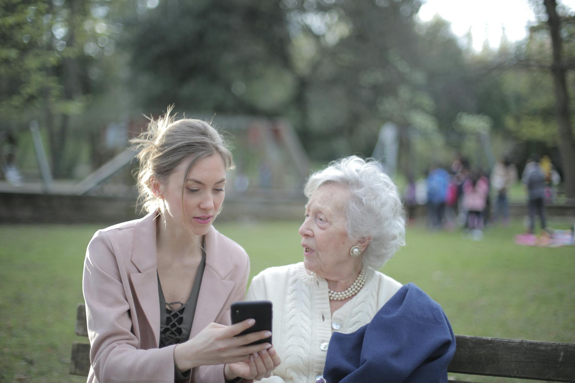 Concentrated female in elegant wear sitting on bench in park and instructing elderly mom on use of cellphone