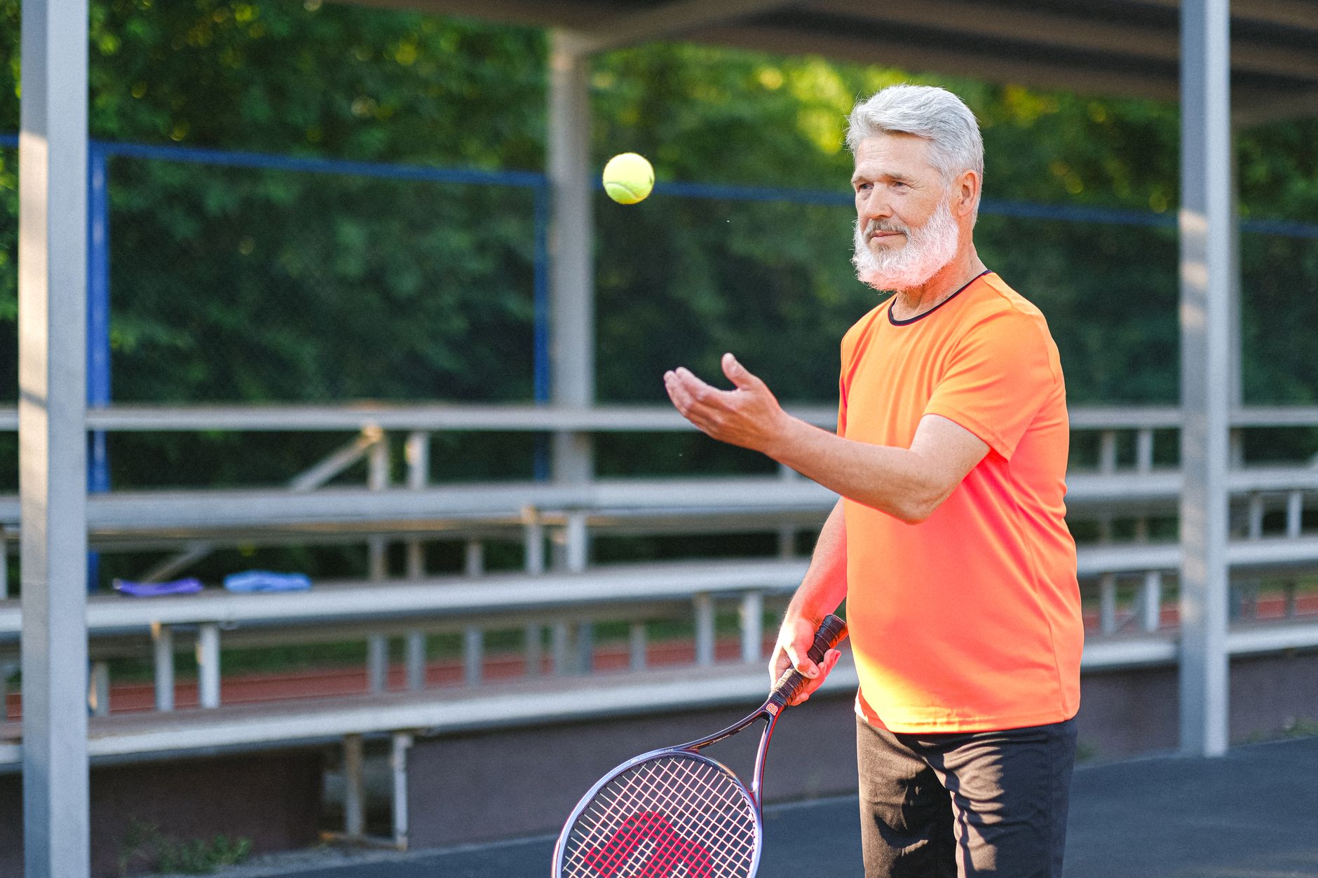 Side view of concentrated senior sportsman with beard in bright sportswear playing tennis on sports court in sunny day