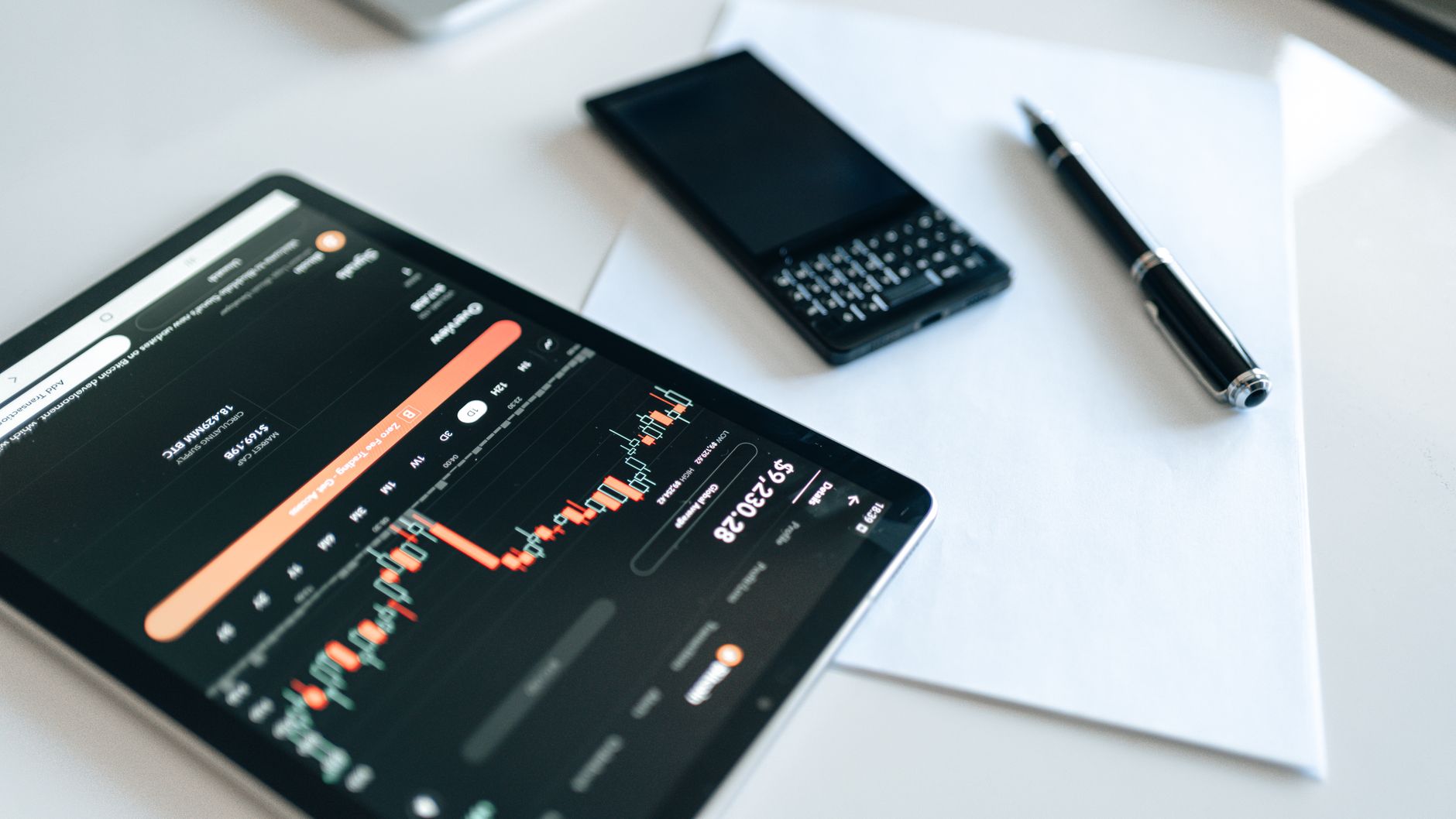 Close-up of a tablet displaying financial charts with a phone and pen on a white surface.