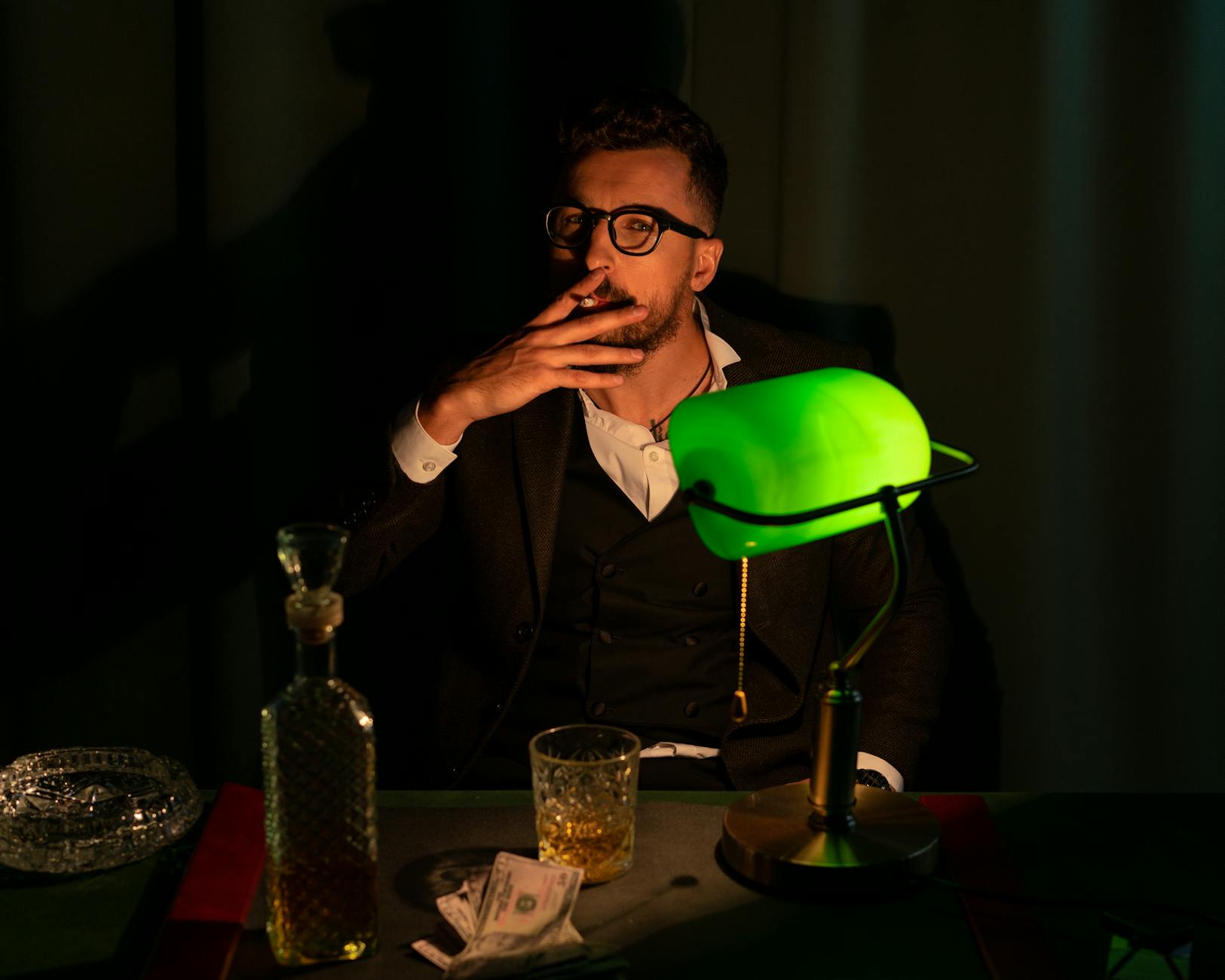 A sophisticated man smoking, seated by a green banker lamp with whiskey and cash, exuding vintage luxury.
