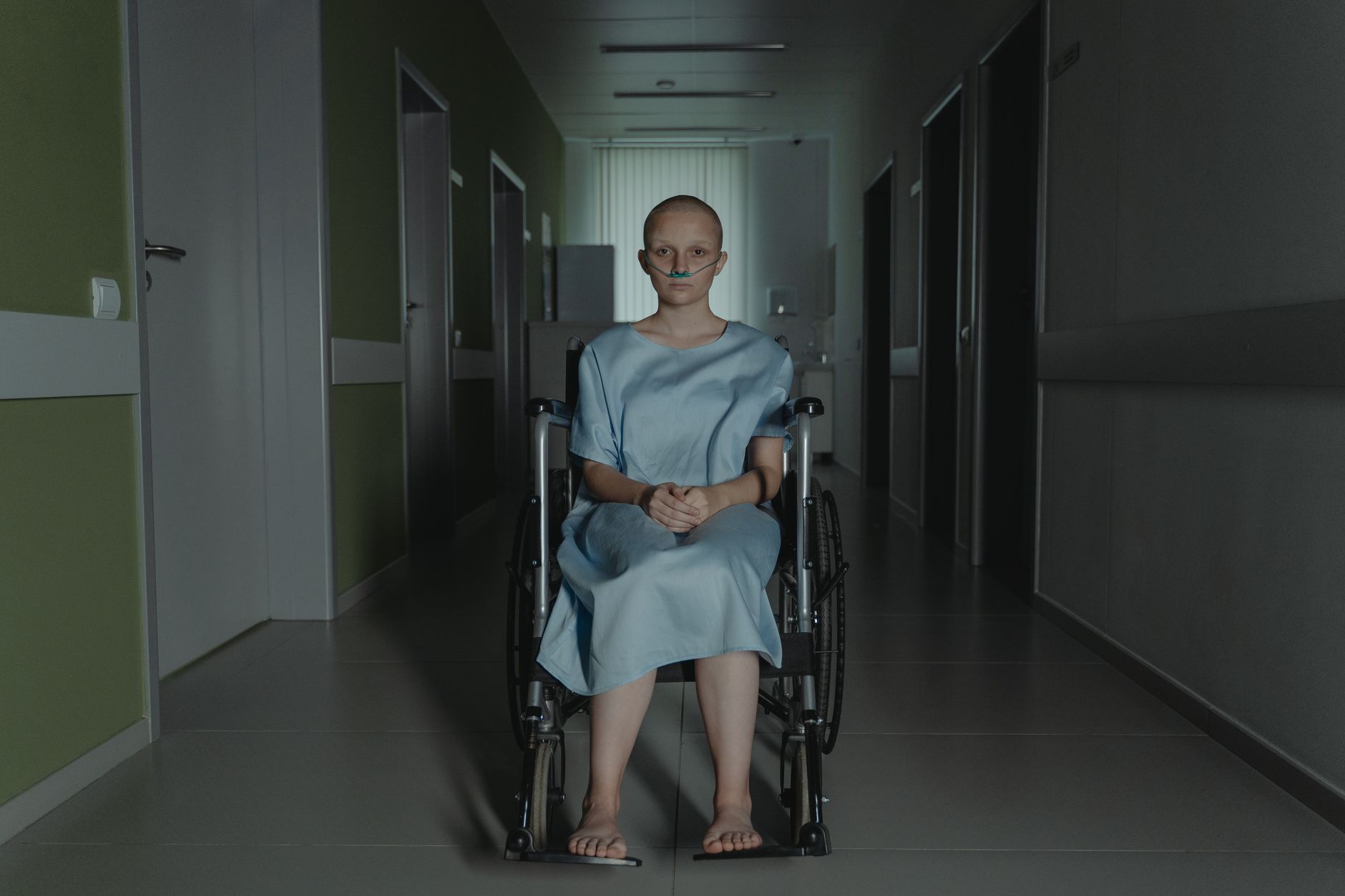 A young woman in a wheelchair sits in a hospital corridor wearing a gown, evoking themes of health and resilience.