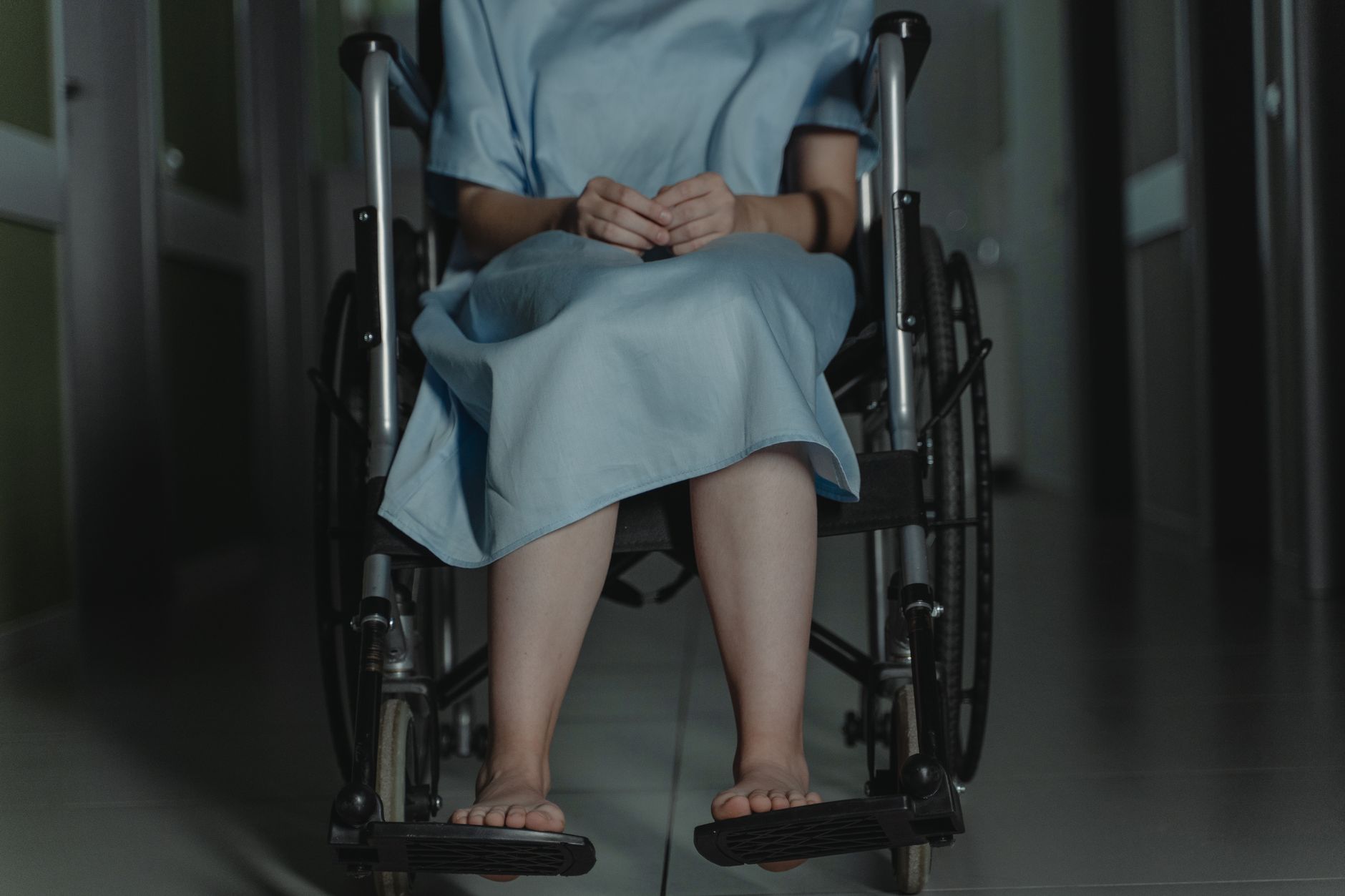 Person sitting barefoot in a wheelchair wearing a hospital gown in a dim hallway.