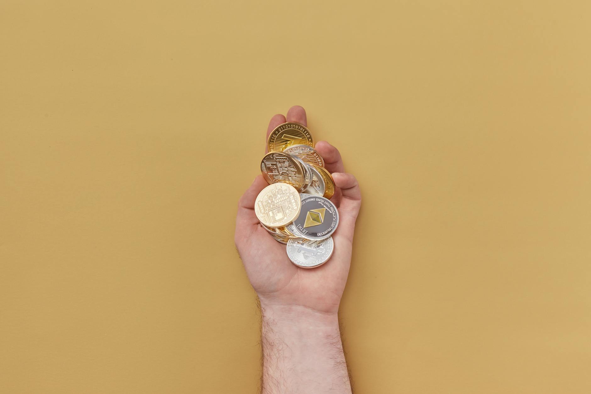 Close-up of a hand holding various cryptocurrency coins against a yellow background.